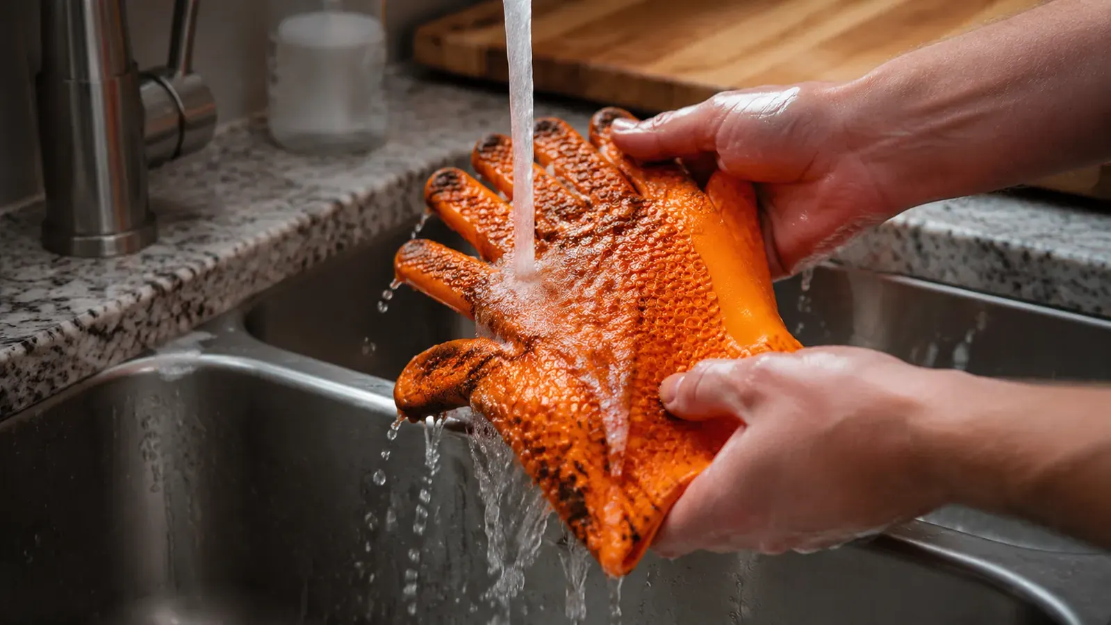 Silicone BBQ gloves being rinsed clean under a kitchen faucet with soap suds visible