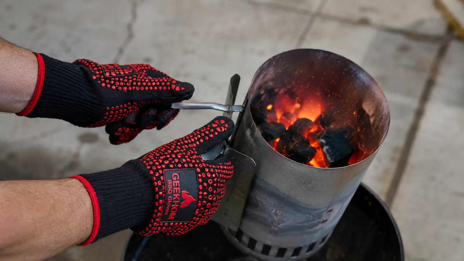 Hands in GEEKHOM silicone gloves gripping a charcoal chimney starter full of glowing coals