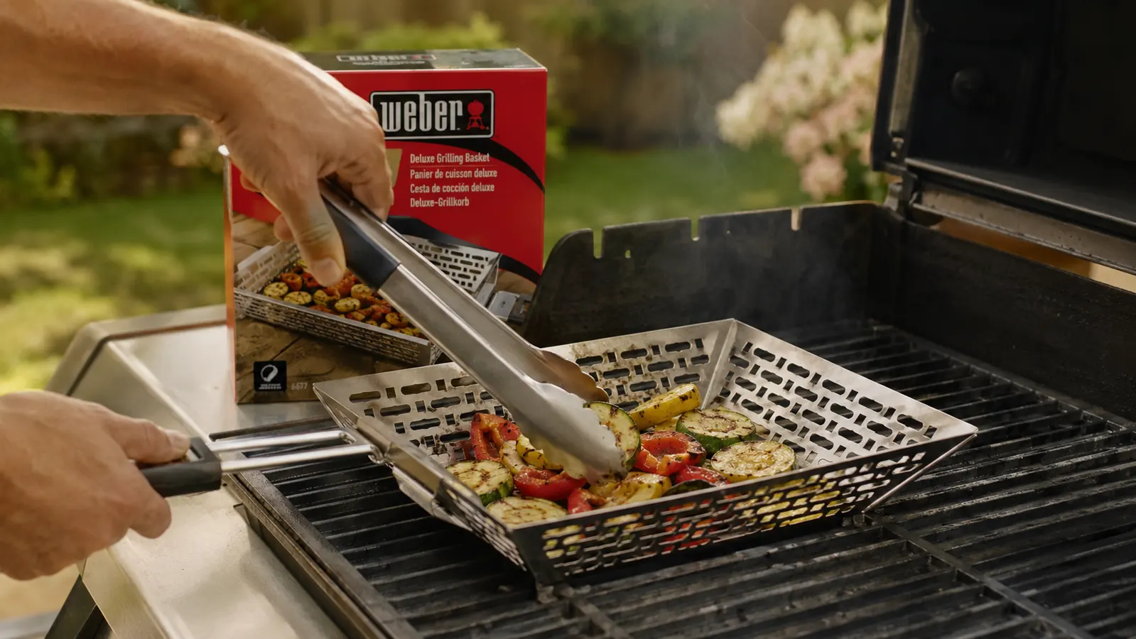 Grilled vegetables with char marks being tossed in a Weber grill basket using long-handled tongs