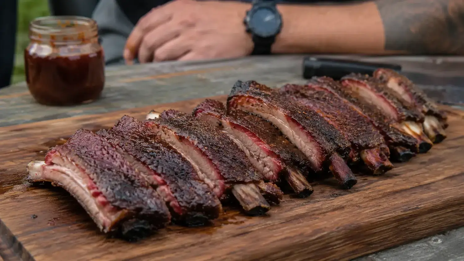 Sliced smoked ribs on a wooden cutting board with a small bowl of BBQ sauce beside them