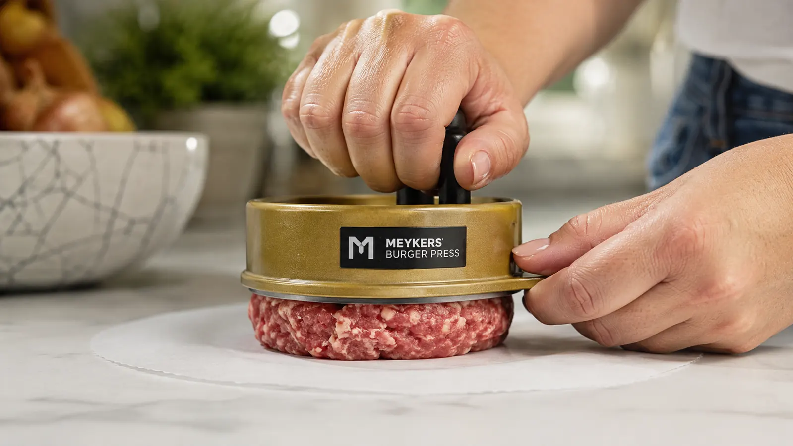 Hand pressing the Meykers burger press down onto a ball of ground beef sitting on a white wax patty paper