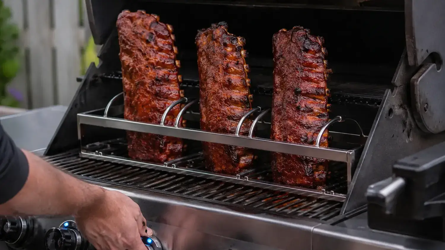 Three full racks of ribs standing upright in a rib rack on a gas grill with the lid open, showing even spacing between each rack